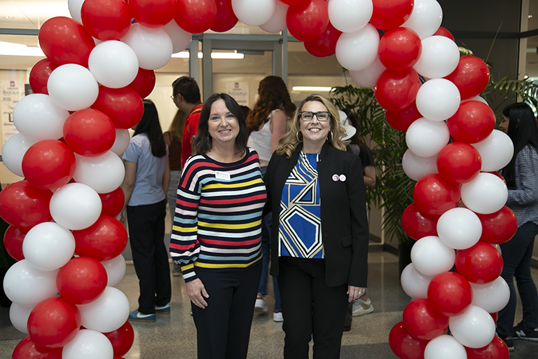 Two people wearing name tags stand together under a red and white balloon arch. There is a large crowd in the background.