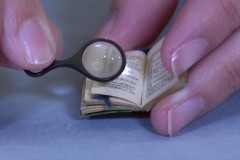 Close-up photo of a person's fingers holding a small magnifying glass and a tiny miniature book.