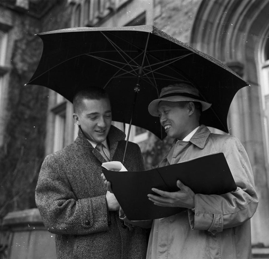 Black and white photograph of students looking at a book under an umbrella.