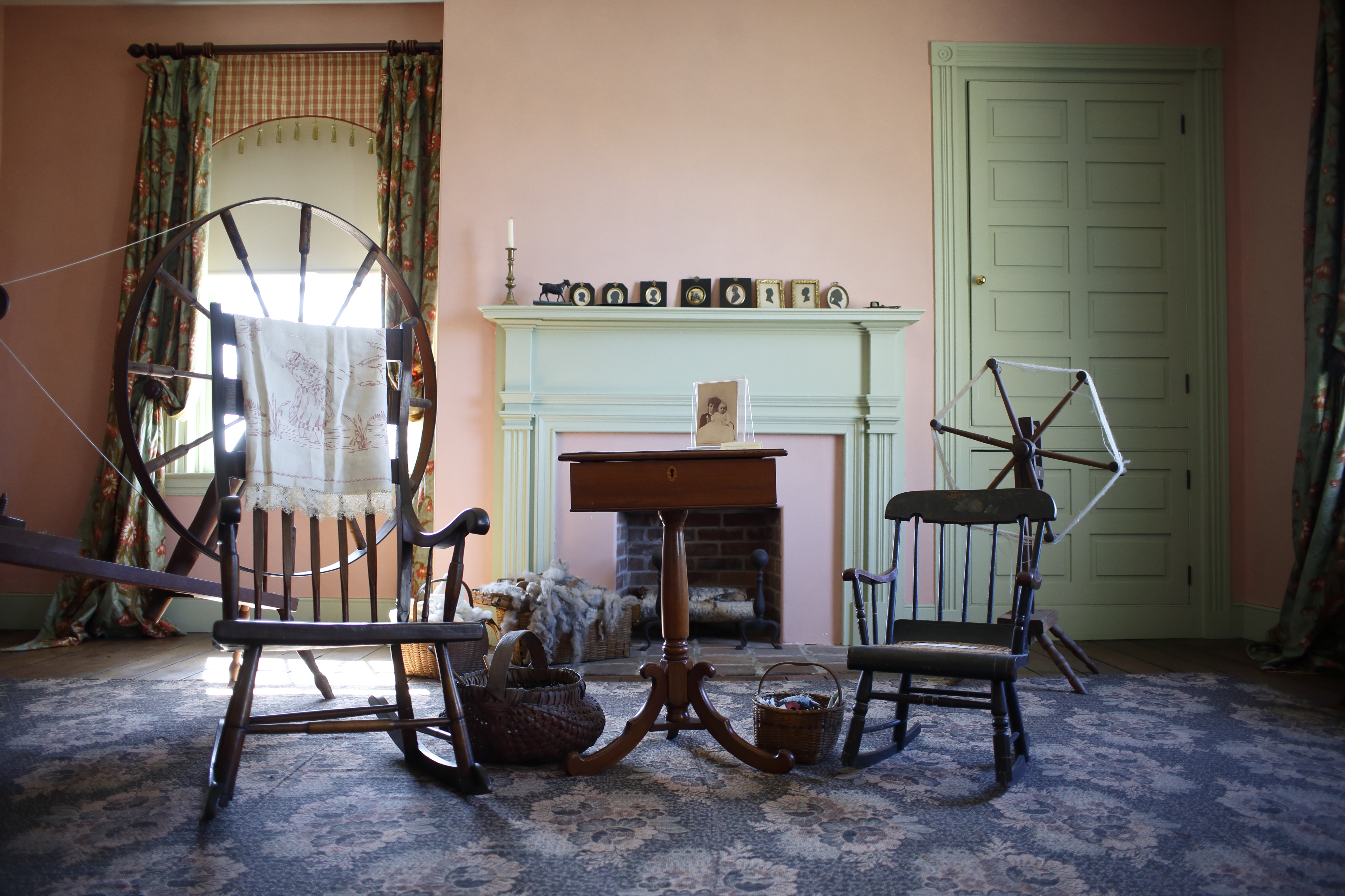 A photo showing empty rocking chairs in front of a fireplace in an historic home. A spinning wheel is visible. 