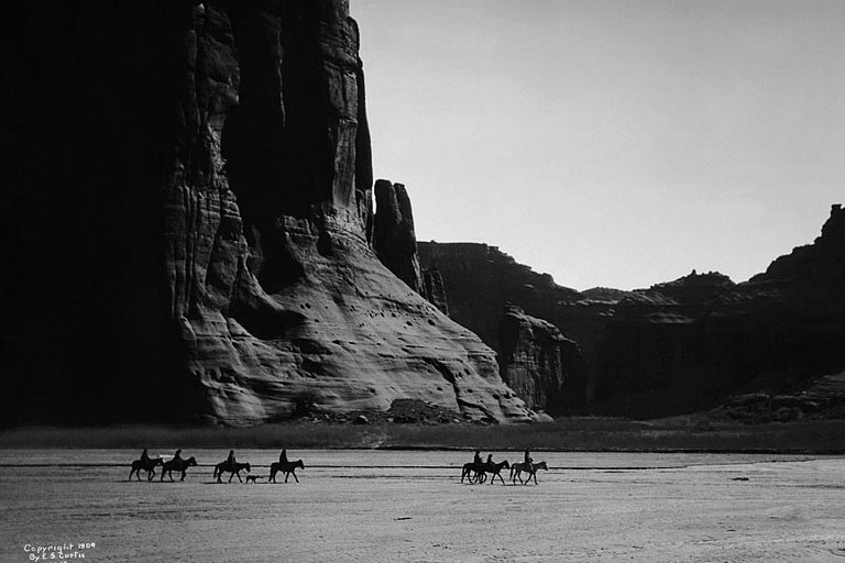 Seven native Navajo ride on horseback with a dog against a background of canyon cliffs.