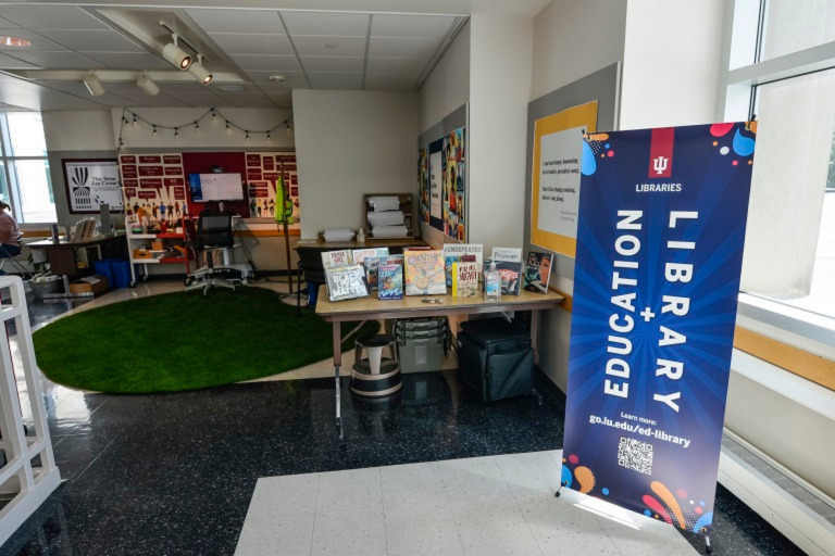 In a room with windows on two sides, there is a large round green rug, a desk with a computer monitor, a table displaying several picture books, a banner that says EDUCATION LIBRARY, and some other things.