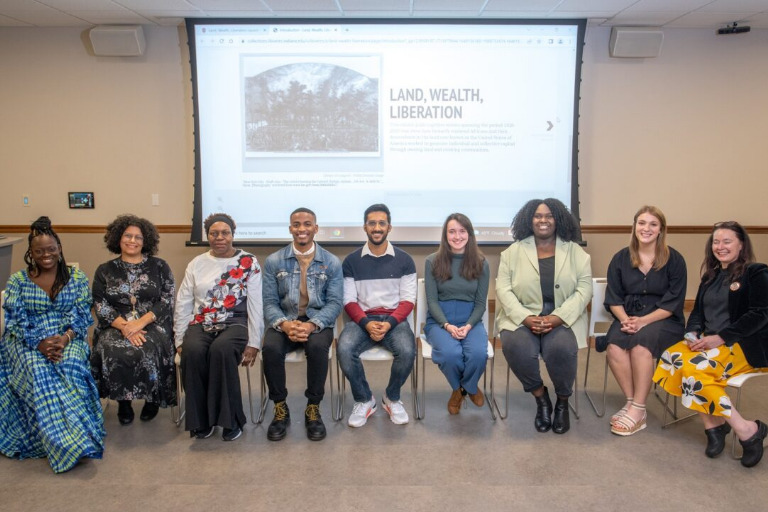 Students and librarians who worked on the Land, Wealth, Liberation project are seated in a row in front of a screen displaying the digital resource. They are smiling with pride in their work.