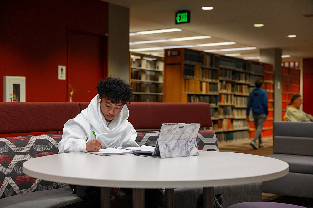 With a laptop nearby and taking notes by hand, a student studies at a library table.