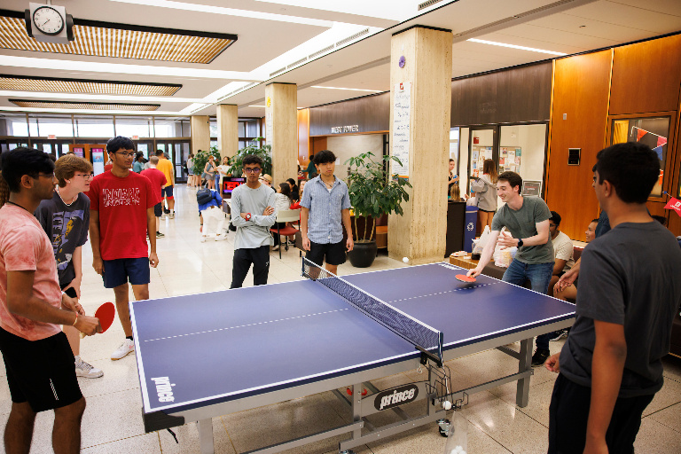 Students play a game of table tennis in the Wells Library lobby while other students watch.