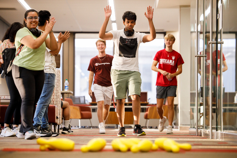 The same student from the previous image raises his hands in triumph as all of the bowling pins have been knocked down. Several students around him smile and applaud.