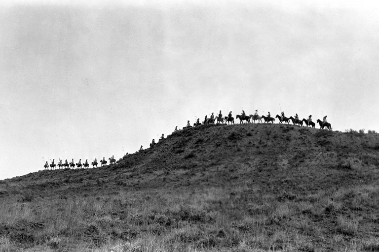 in a black and white image a line of people sitting on horses traverses a hilltop 