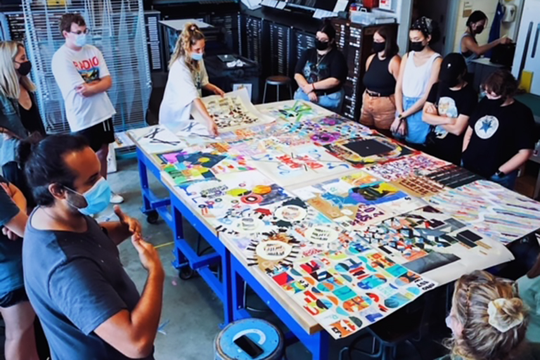 A table full of colorful art is a the center of a group of college students looking a an instructor who speaks with his hands up. All wear masks.