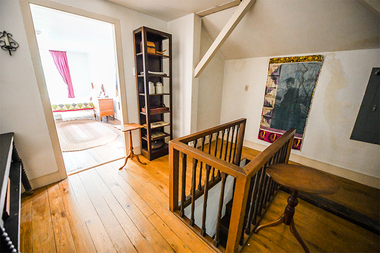 A room with low ceiling and wooden floor is pictured with a staircase railing in its center
