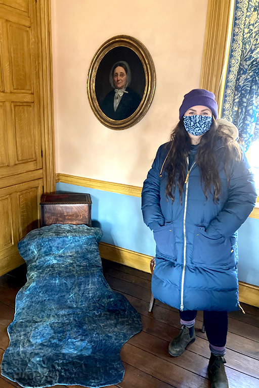 A person in a blue coat stands next to a blue quilt draped over an antique chair.