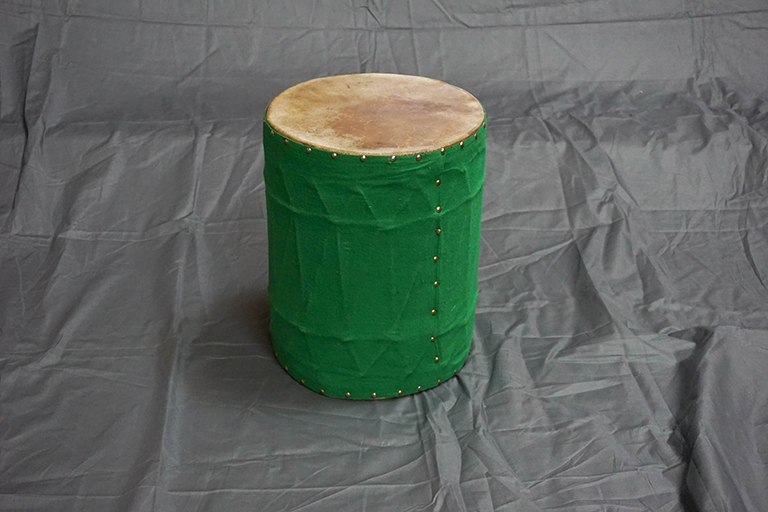 A cylindrical barrel covered in green cloth and placed on one end is displayed on a grey cloth surface.