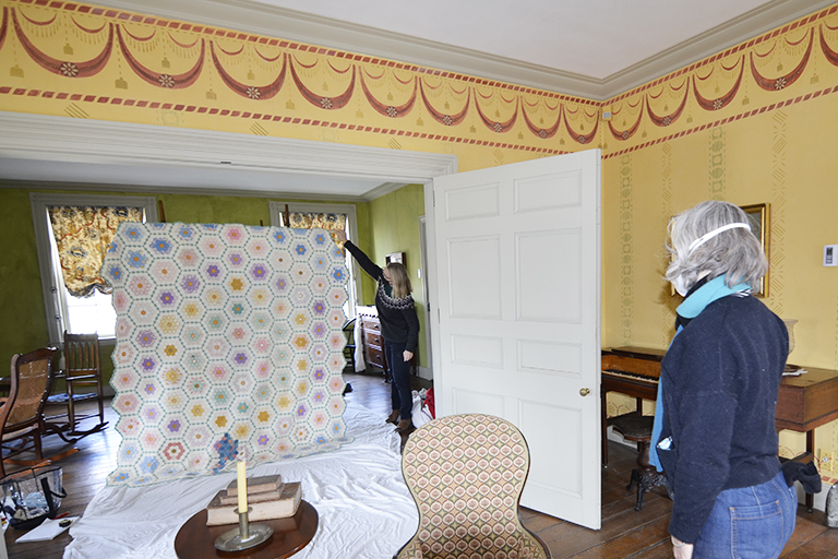 A large quilt is taller than the person holding it so the artist can see it fully upright inside a historic house museum.