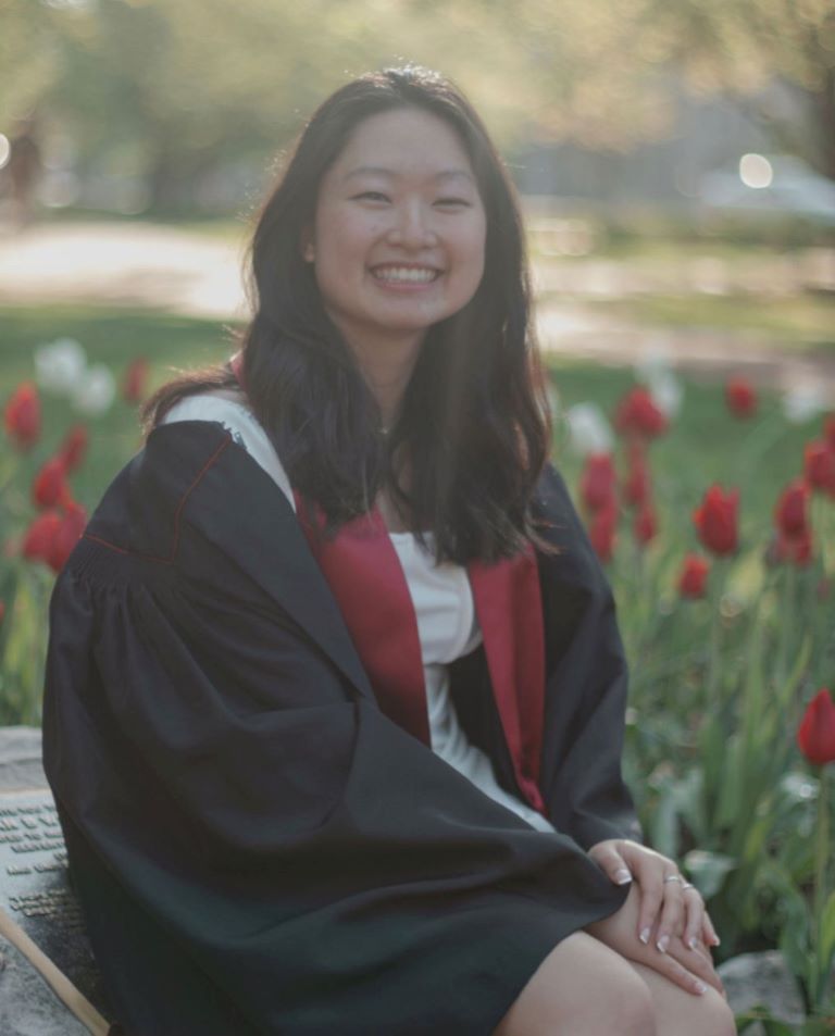 This is a graduation portrait that appears to be taken in the morning when the light is soft. Christina, who is wearing a white dress, is in her graduation gown with its crimson scarf. She looks very happy.