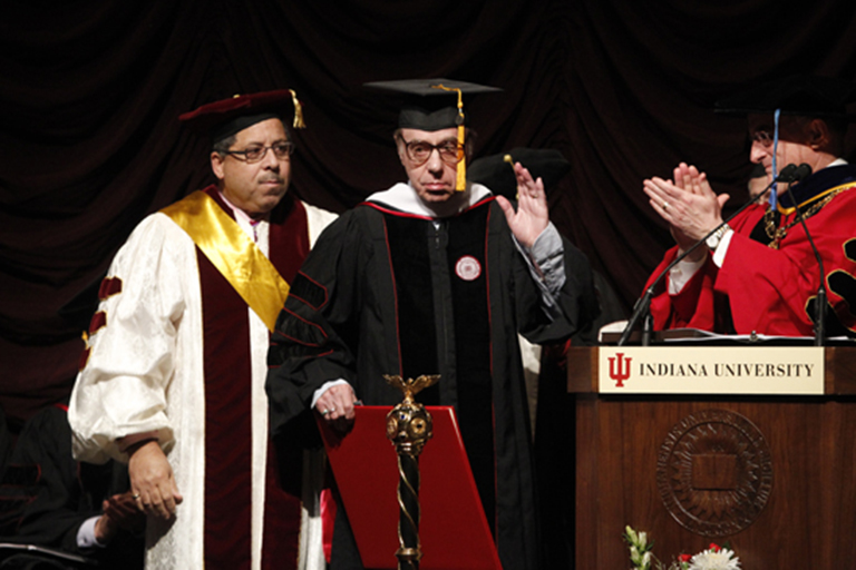 Two-time Academy Award nominee Peter Bogdanovich acknowledges the audience inside the IU Cinema upon receiving an honorary degree from President Michael McRobbie in 2011. Three individuals are in the photo, all wearing full regalia and standing at a podium.