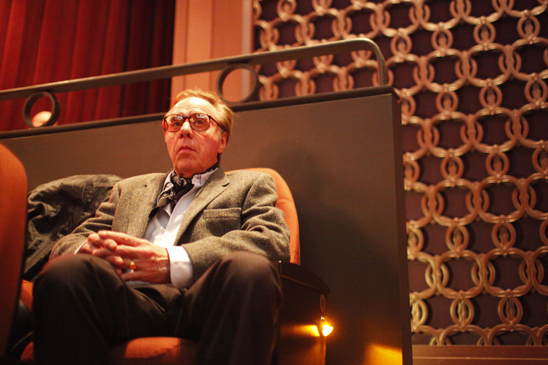 A man wearing glasses, suit, and tie is seated in a theater.