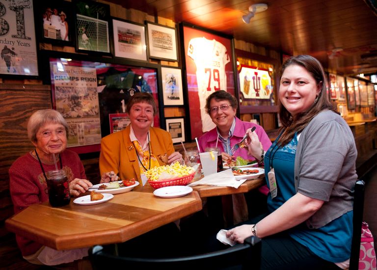 Four generations of Jay women sit at a table eating: the oldest is small and in red; the two in the middle are beaming, one in yellow and the other in pink; and the youngest is in the forefront in gray and blue.