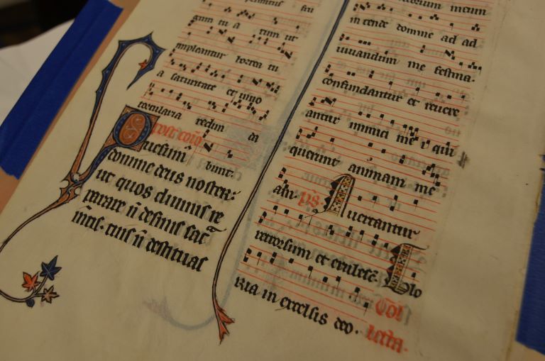 A medieval musical script with red, blue, and yellow flowers in the corner. The text is Latin.