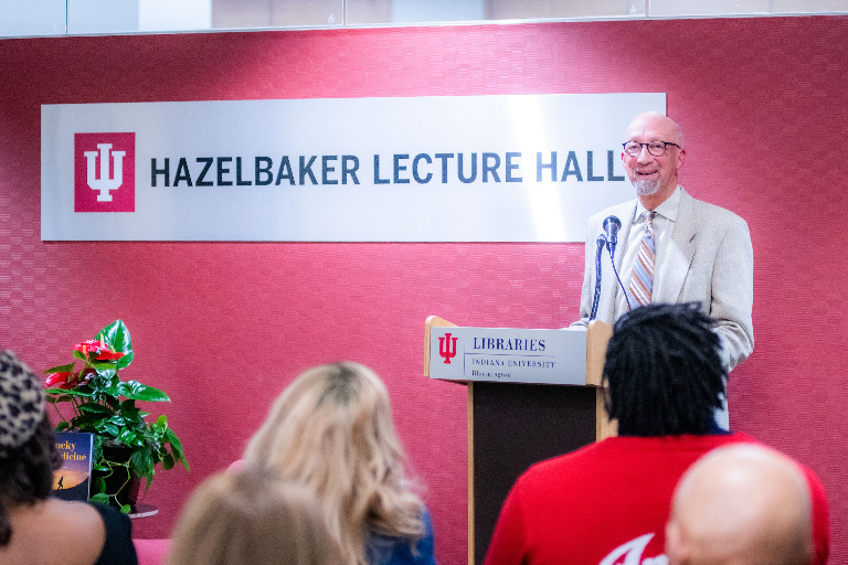 A man in a light-colored suit and tie stands at a lectern. Behind him a sign reads "Hazelbaker Lecture Hall."