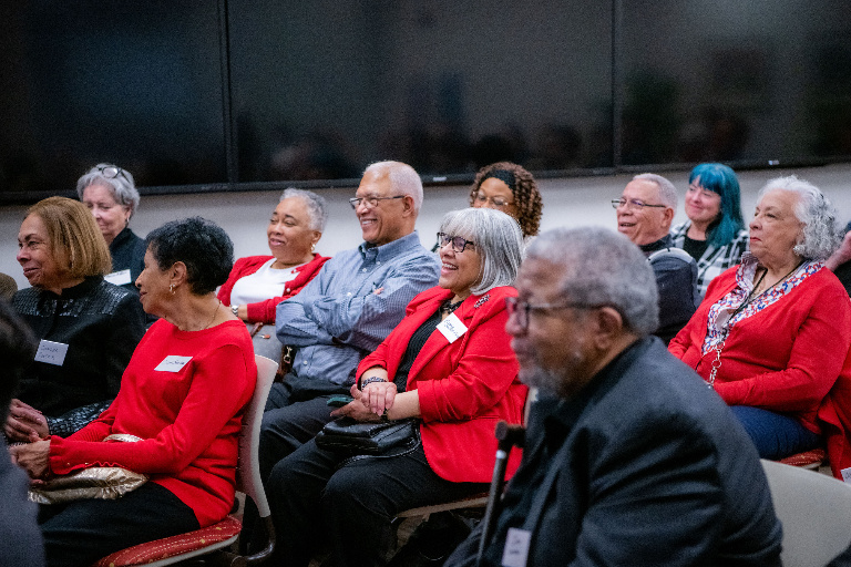 Members of the audience, many wearing red tops or jackets, listening. Several are smiling or laughing.