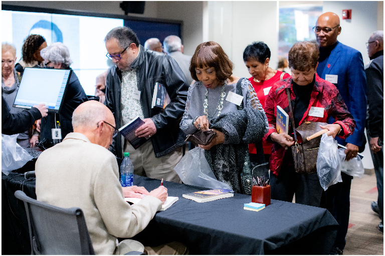 A man seated at a table, his back to the camera, signing books. An eager crowd waits with books in hand to be signed.