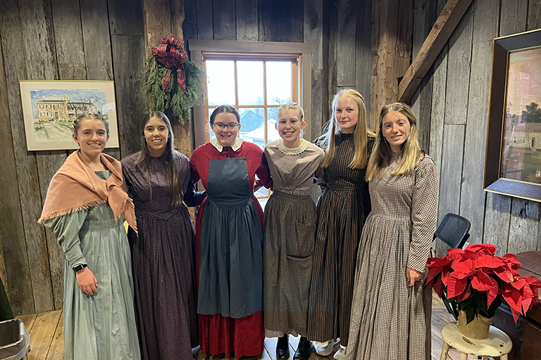 Six young adults stand together smiling. They are wearing period attire representing females in 19th century America