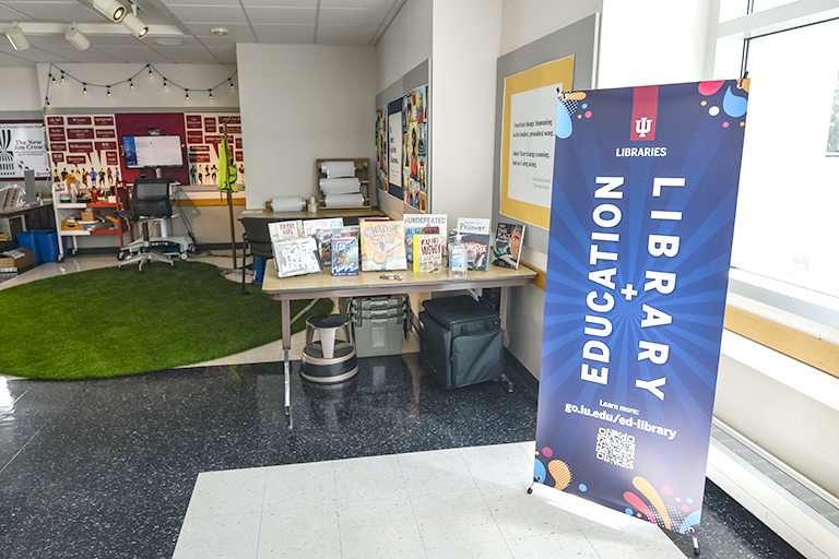 A light filled open space is arranged with a welcome table and tall banner indicating this is a place for the Education Library.  In the background are many carts full of books, a green indoor rug and walls full of posters and fliers.