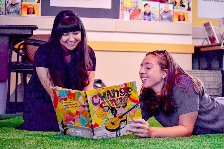 Two young adults sprawl across green indoor carpeting as they smile and look at a colorful children's picture book. The visible title reads, "Change Sings"