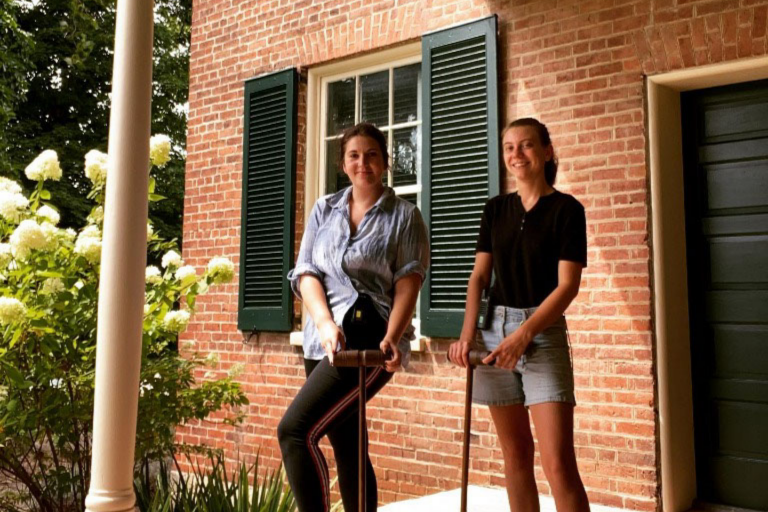 Two college-aged women standing on porch of brick home holding croquet mallets/