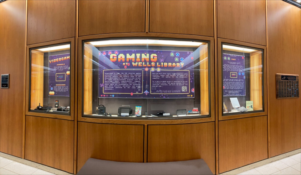 A photo of a large poster display. It's titled "Gaming at Wells Library," and has a bright, retro pixel theme. At the bottom of the case, several gaming consoles are displayed.