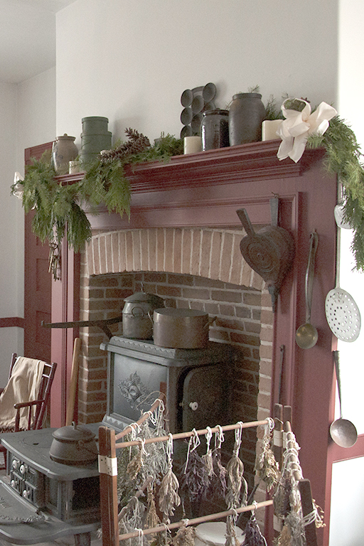A live greenery garland is draped across the high wooden mantel of an 1840's cooking hearth