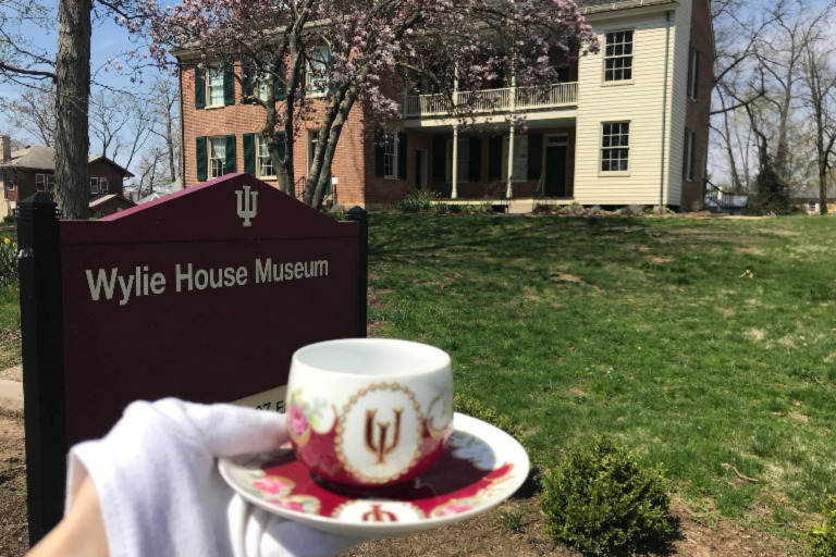 An ornate porcelain IU cup and saucer are held by a white-gloved hand on the lawn of the Wylie House Museum. The front of the house can be seen in the background.