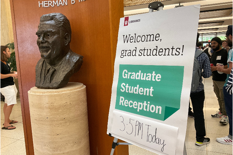 The bust of Herman B Wells in the library lobby, and a sign on an easel reading "Welcome, grad students! Graduate Student Reception 3-5 PM Today"