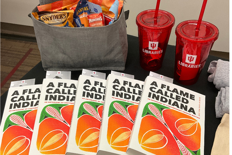 Several copies of the book "A Flame Called Indiana," a totebag filled with packaged snacks, and red plastic IU Libraries tumblers.