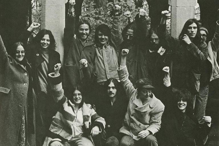 Group portrait of students standing and sitting in front of an archway in 1970s era clothing, each with a fist raised in the au.