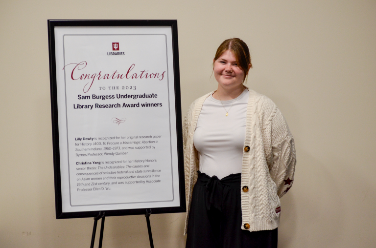 Lilly stands next to a large congratulatory sign. She is in elegant black and white. Her red hair is pulled back and her smile is proud.