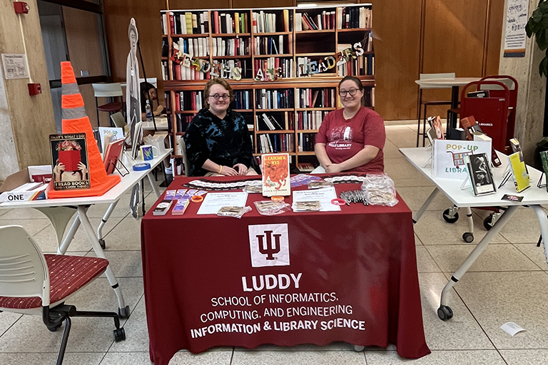 Two people sit behind a table surrounded by books. The tablecloth has an IU logo on it and the name Luddy. 