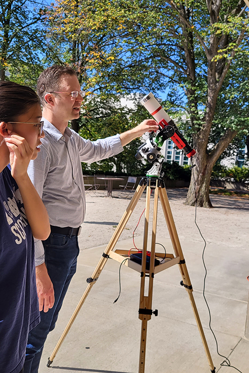 Two people stand outside examining a telescope pointed at the ground