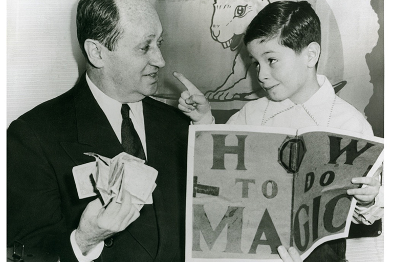 An older person wearing a shirt, tie and jacket looks to a younger person with short hair and a button up shirt.  A poster in view reads, how to do magic