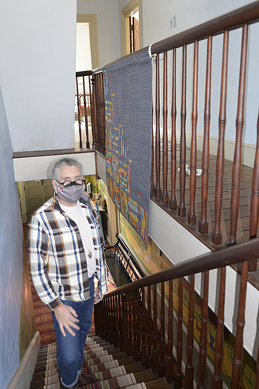A person wearing glasses and a flannel shirt stands on a staircase with a quilt on the railing behind him.