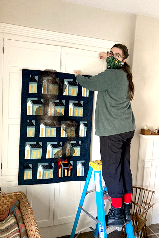 An artist stands on a blue ladder to hang a dark colored quilt on a white wall. The quilit squares features houses and one is burning.