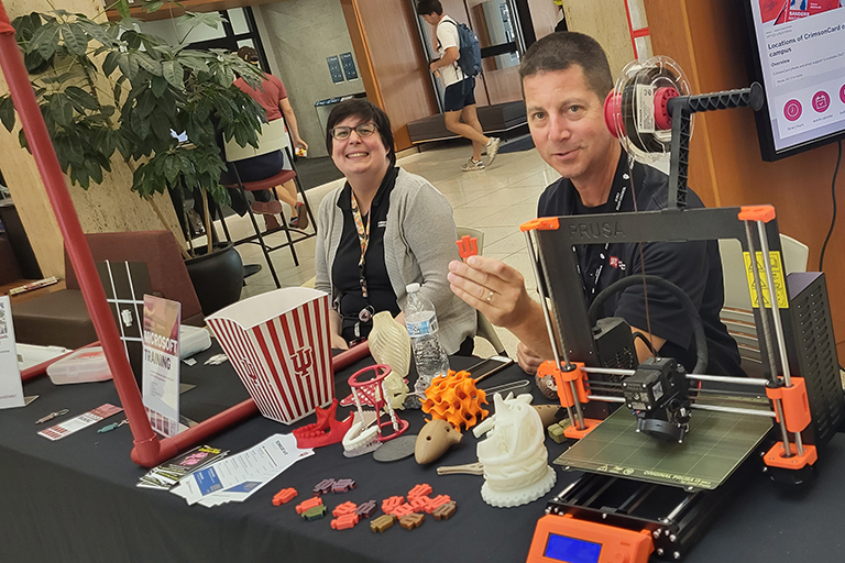 Two people are smiling and seated behind a table full of supplies for a 3D printer
