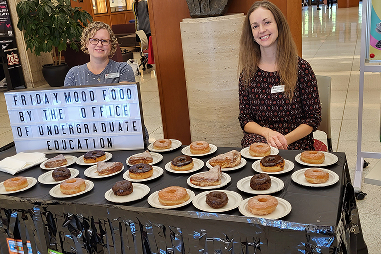 Two people wearing nametags sit behind a table filled with donuts on small plates and a sign on the table reads Friday Mood Food with the office of Undergraduate Education