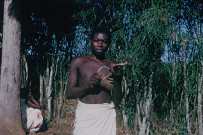 A boy stands amidst vegetation holding a small musical instrument.