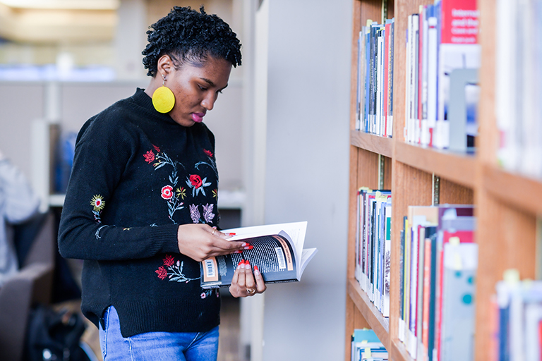 A woman in a black sweater and bold earrings browses a bookshelf in the library