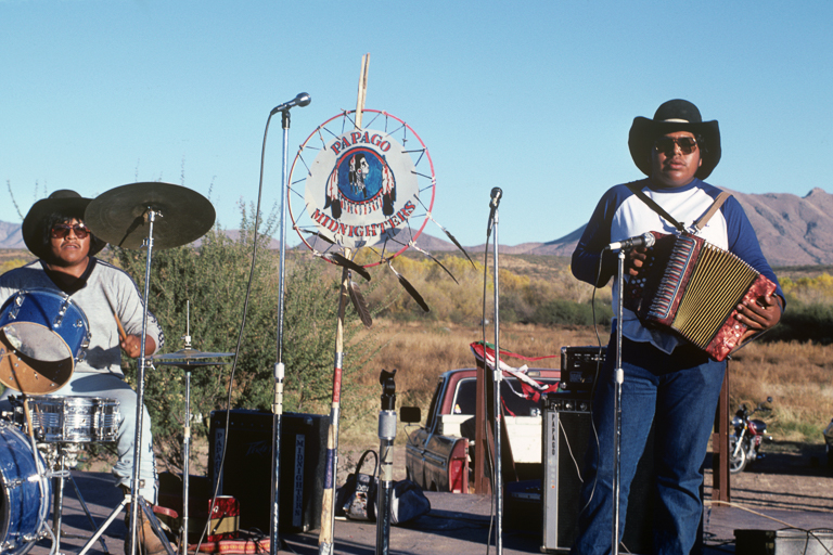 A band with drums, accordian, and electric guitar performs outside in front of desert mountains.
