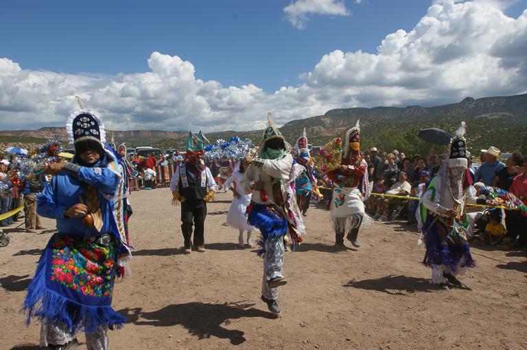 Costumed people dance outside under a big blue sky with white clouds and surrounded by desert mountains and spectators.