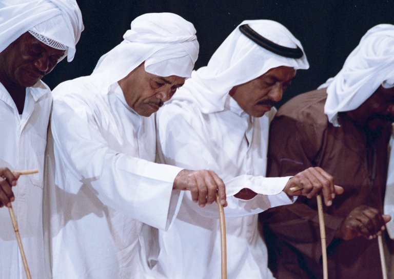Two men in white robes and head coverings stand with other men in a line and look down while leaning on staffs.