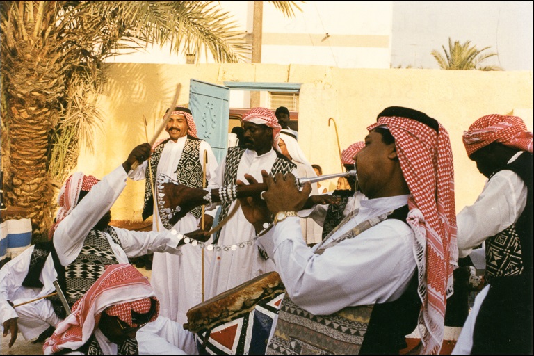 Men in red-checked head coverings and black vests play trumpets, drums, and other instruments near a clay wall and a palm tree.