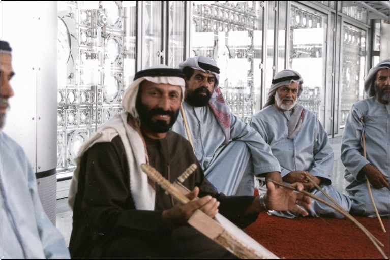A musician in a dark robe and white head covering sits with other men on a red rug. he holds a wooden instrument and smiles at the camera.