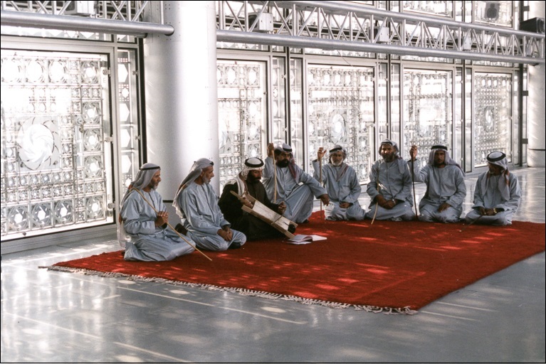 Seven men in mostly grey robes and head coverings kneel along two edges of a red rug in an ornate grey room and watch an eighth man in a dark robe play an instrument.